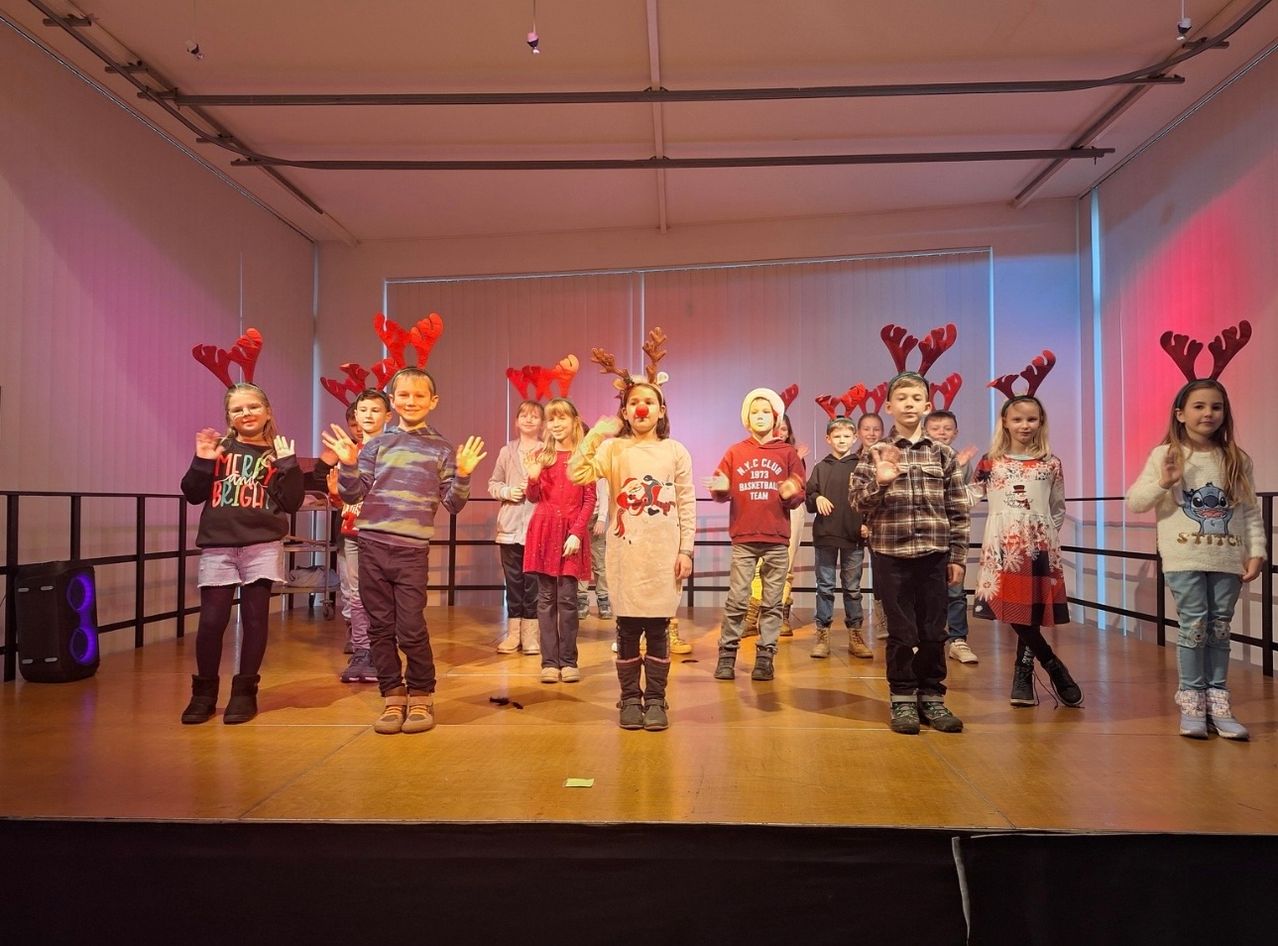 Eine Gruppe von Kindern mit roten Rentier-Kopfbändern und festlicher Kleidung steht auf der Bühne und singt oder spielt wahrscheinlich ein Weihnachtslied oder Theaterstück. Der Hintergrund ist eine Mischung aus rosa und blauen Farbtönen, wobei einige Kinder Turnschuhe und andere Stiefel tragen.