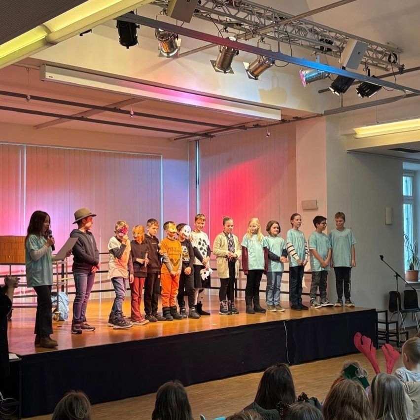 Kinder stehen auf der Bühne, tragen Kostüme, und eine Frau hält ein Mikrofon. Hinter ihnen beleuchten rosa und rote Lichter die Wand. Das Publikum beobachtet von unten.