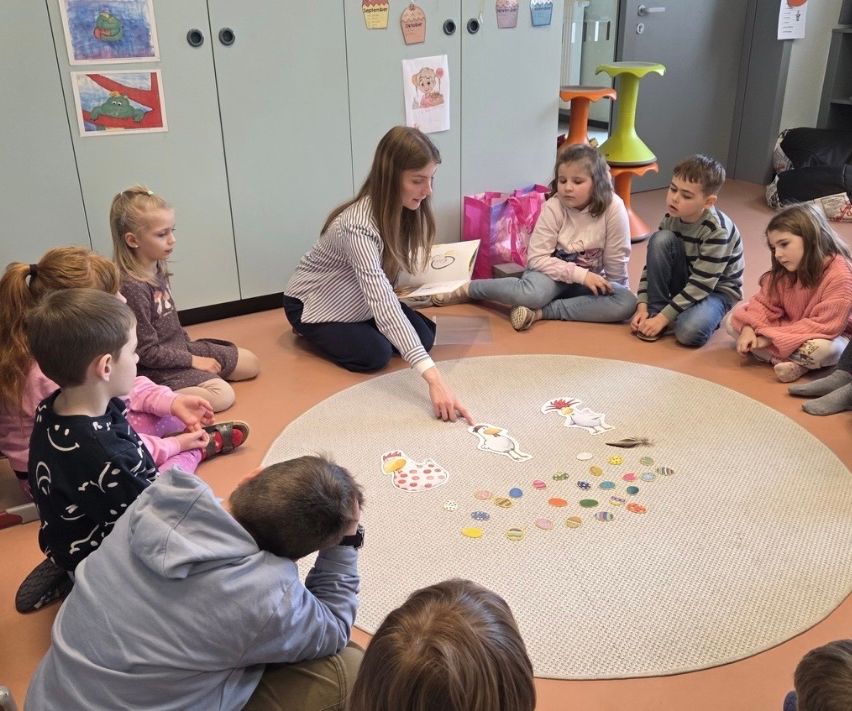 Szene im Klassenzimmer, in dem eine Lehrerin Kindern, die im Kreis sitzen, aus einem Buch vorliest, ein Spielteppich mit Spielsteinen und Kunstwerke an den Wänden.