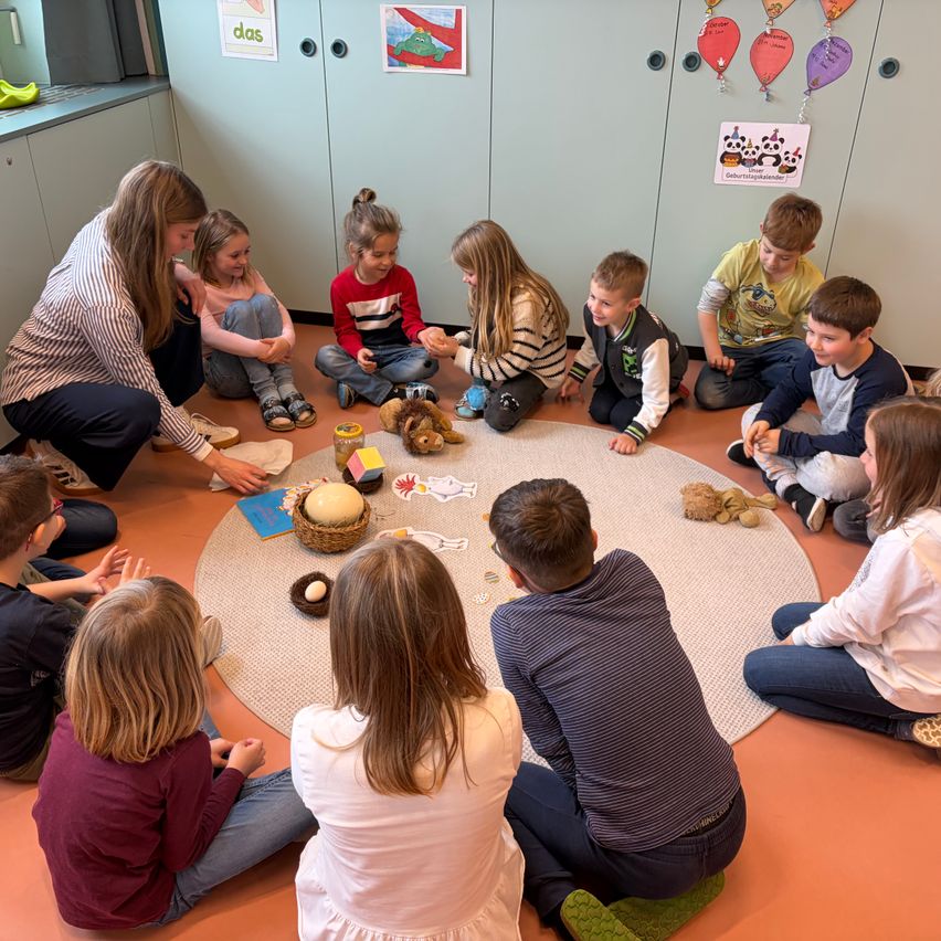 Eine Gruppe von Kindern sitzt in einem Kreis um eine Frau herum und beschäftigt sich mit Spielzeug und Bastelarbeiten auf dem Boden. Der Raum ist mit bunten Dekorationen geschmückt.