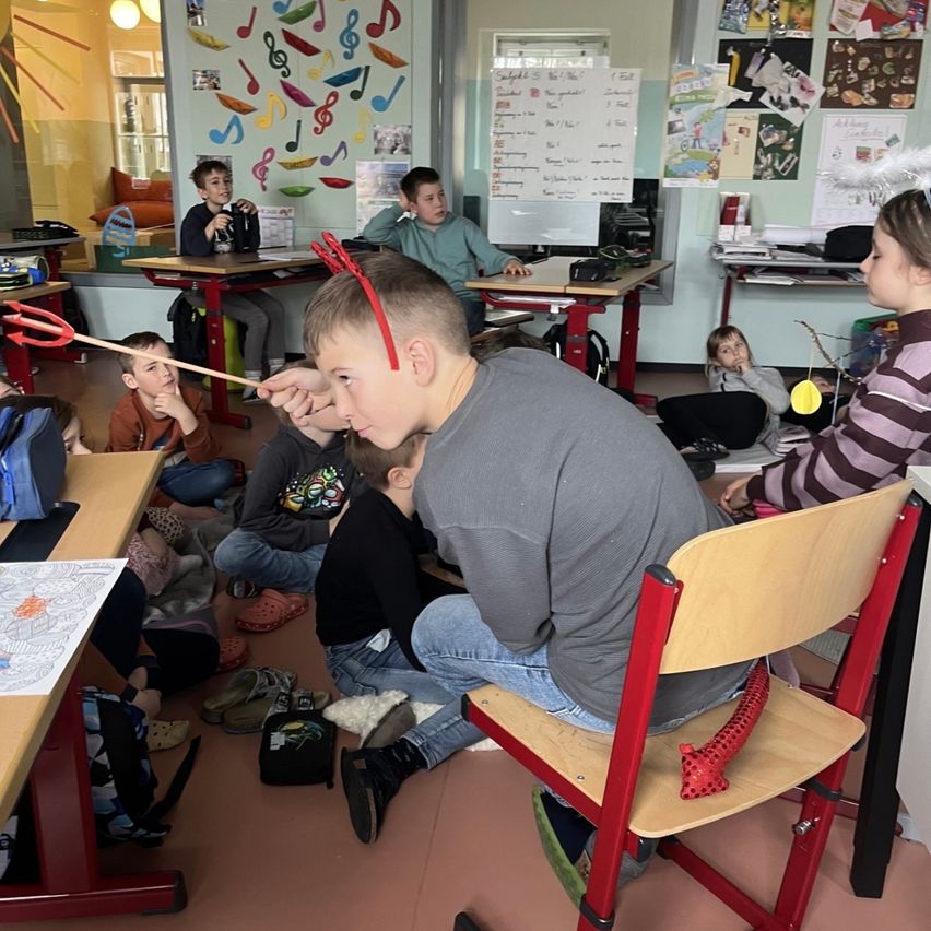 Ein Klassenzimmer mit Schülern, die in Stühlen und auf dem Boden sitzen. Ein Junge mit roten Teufelshörnern schaut sich etwas an, während andere um ihn herum sitzen. Es gibt Schreibtische, eine Tafel und Dekorationen an den Wänden.