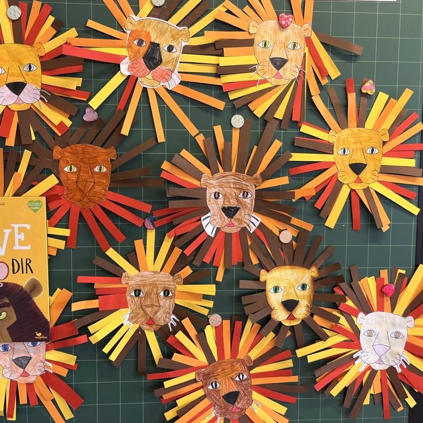 Eine Sammlung von Papierlöwenkopf-Basteleien, die in einem Kreis auf einem grünen Hintergrund angeordnet sind. Jeder Löwenkopf hat unterschiedliche Gesichtszüge und ist von mehrfarbigen Papierstreifen umgeben.