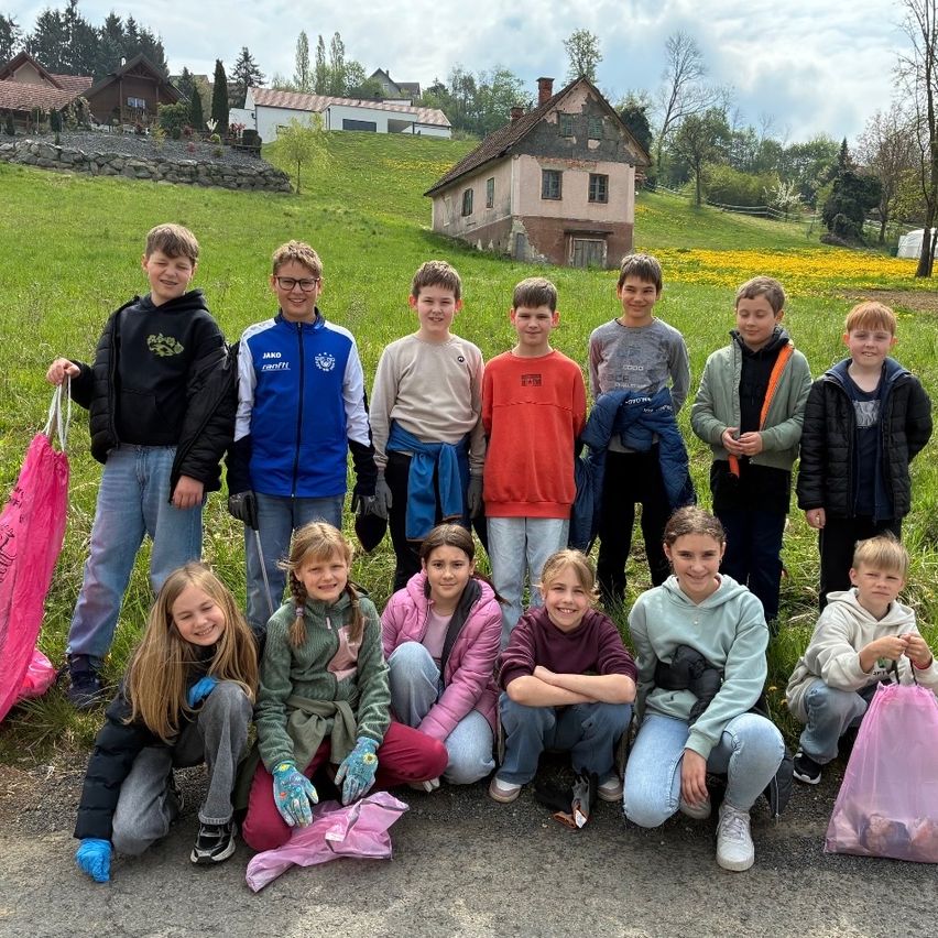 Eine Gruppe von Kindern steht und hockt in einem Feld und hält Müllsäcke. Hinter ihnen sind Häuser und ein großes grünes Feld.
