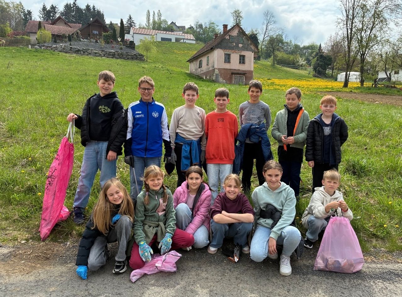 Eine Gruppe von Kindern steht und hockt in einem Feld und hält Müllsäcke. Hinter ihnen sind Häuser und ein großes grünes Feld.