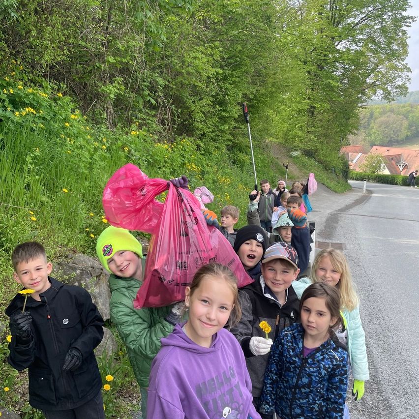 Eine Gruppe von Kindern mit pinken Mülltüten, die für ein Foto an einer Straßenseite lächeln. Sie sind von grüner Vegetation und Blumen umgeben.