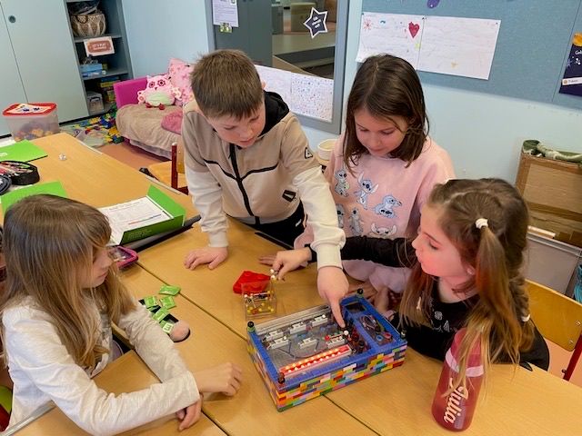 Vier Kinder arbeiten gemeinsam an einem farbenfrohen elektronischen Projekt aus LEGO, das auf einem Tisch in einem Klassenzimmer aufgebaut ist.