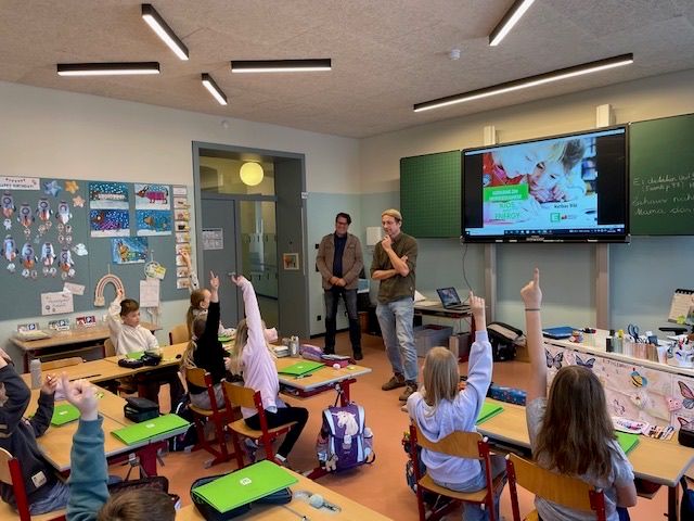 Ein Klassenzimmer mit Schülern, die die Hände heben, zwei Männern, die stehen, einem Monitor an der Wand und farbenfrohen Dekorationen an den Wänden.