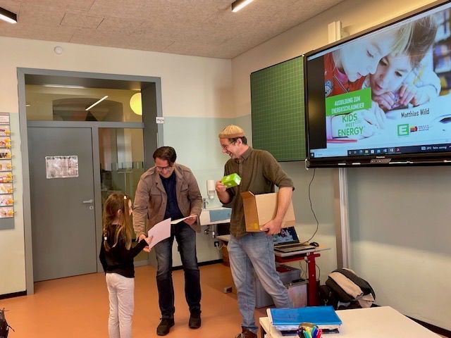 Drei Personen stehen in einem Klassenzimmer, zwei Männer und ein junges Mädchen. Sie halten Papiere und Kisten. Das Mädchen schaut den Mann an, der eine Kiste hält. Ein Monitor zeigt ein Poster mit dem Text Kids Meet Energy.
