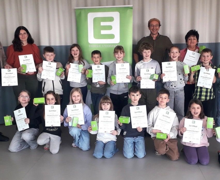 Gruppenfoto von Kindern, die Zertifikate halten und vor einem grünen Banner mit Text und Logos lächeln.