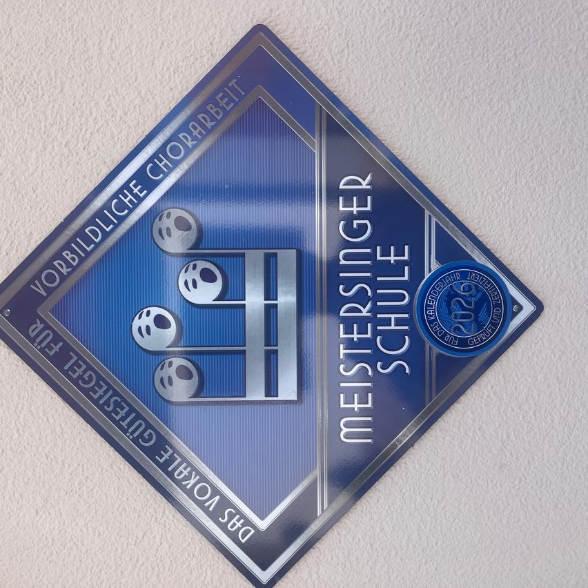 Ein blaues rautenförmiges Schild mit 'Meistersinger Schule' darauf. Es enthält ein Logo und den Text 'Vorbildliche Chorarbeit'. Das Jahr '2024' ist angezeigt.