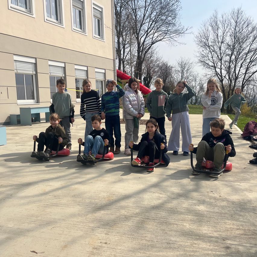 Eine Gruppe von Kindern und Jugendlichen sitzt auf roten Schlitten im Schulhof, einige tragen Jacken und Turnschuhe. Ein Gebäude mit Glasfenstern steht hinter ihnen, und es gibt Bäume und einen roten Schlitten im Hintergrund.