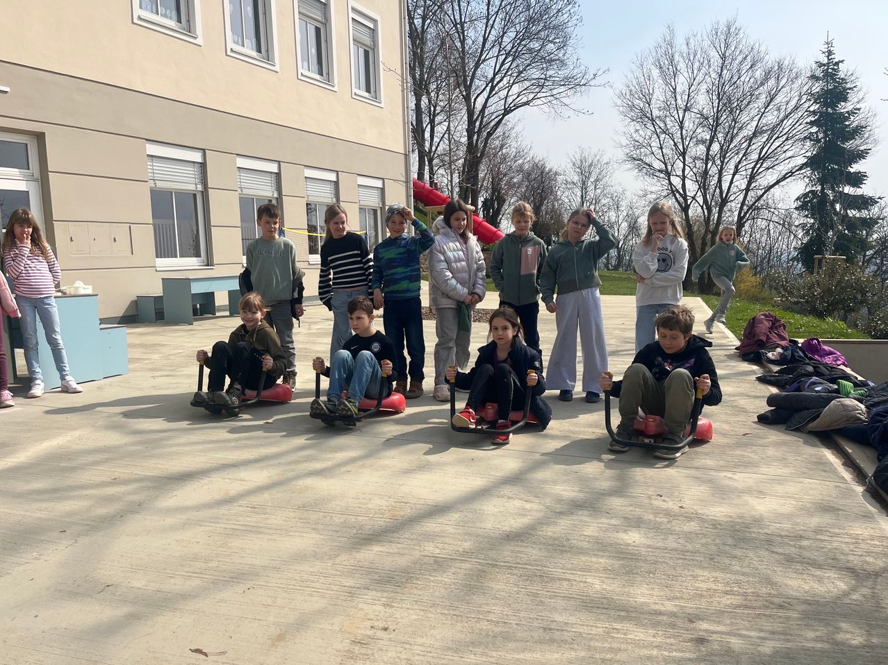 Eine Gruppe von Kindern und Jugendlichen sitzt auf roten Schlitten im Schulhof, einige tragen Jacken und Turnschuhe. Ein Gebäude mit Glasfenstern steht hinter ihnen, und es gibt Bäume und einen roten Schlitten im Hintergrund.