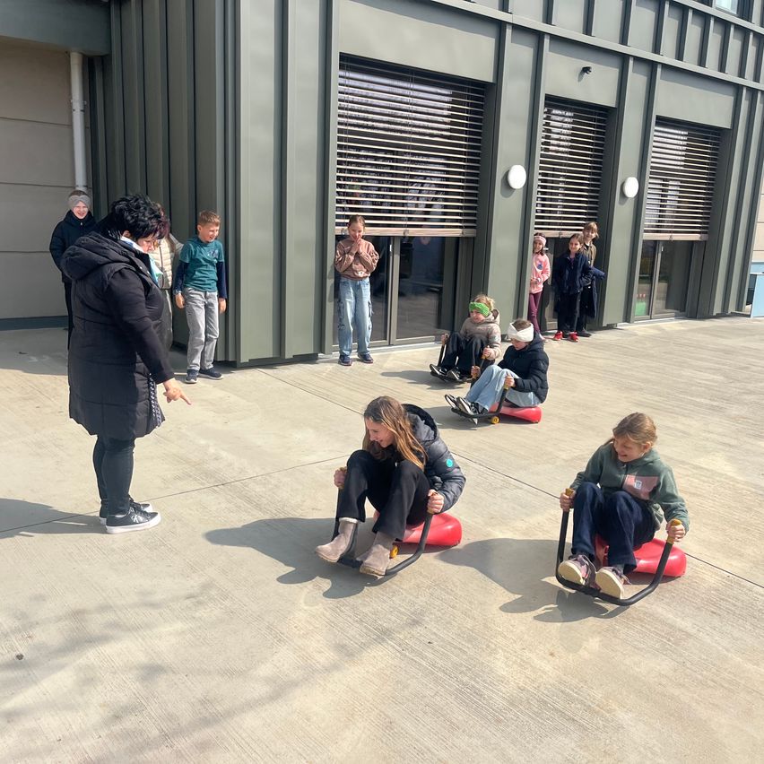 Eine Frau zeigt auf eine Gruppe Kinder auf Schlitten, während ein Gebäude mit Fenstern und Türen hinter ihnen steht.