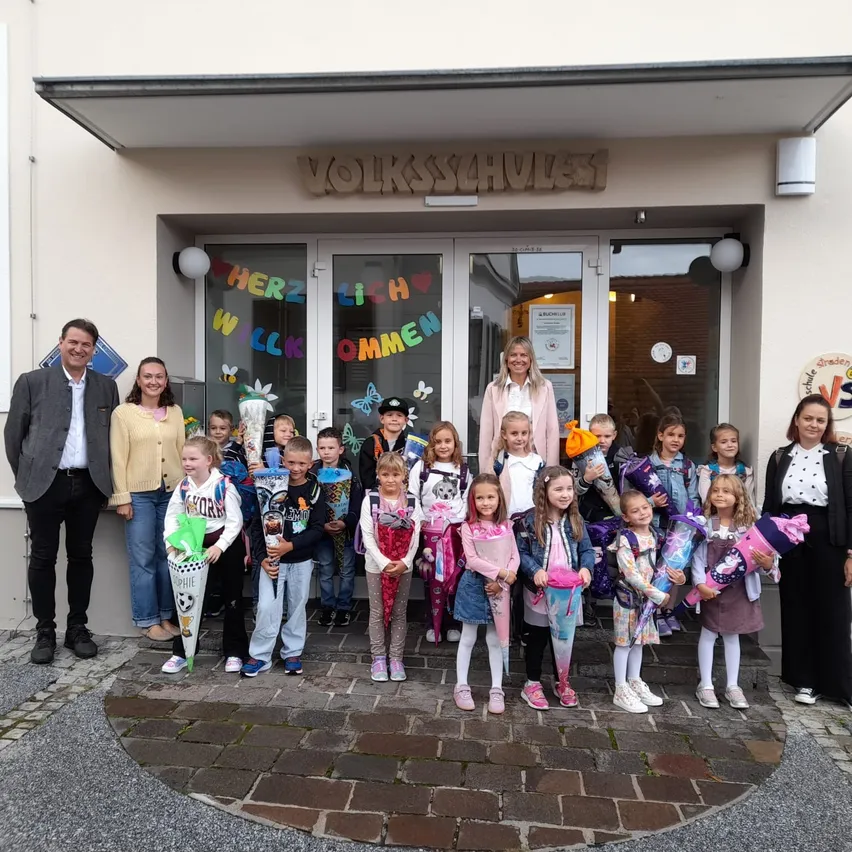Eine Gruppe von Kindern und Erwachsenen steht vor einem Gebäude mit der Aufschrift 'Volksschule'. Die Kinder halten bunte Kegel und Geschenke. Der Eingang des Gebäudes ist mit bunten Buchstaben und Schmetterlingen geschmückt.