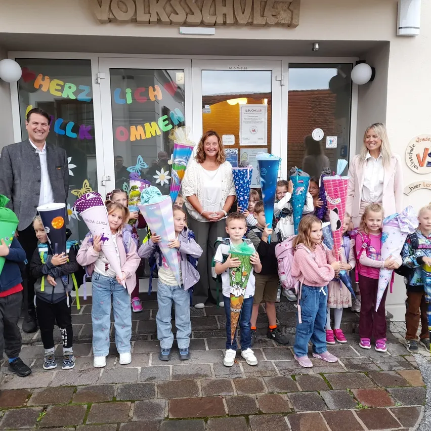 Eine Gruppe von Kindern und Erwachsenen steht vor einem Gebäude, hält bunte Kegel und lächelt. Das Gebäude hat ein Schild mit der Aufschrift 'Herzlich willkommen'.