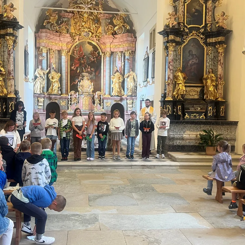 Eine Gruppe von Kindern und Erwachsenen steht in einer Kirche, vor dem Altar mit goldenen Statuen. Einige Kinder halten Papiere.