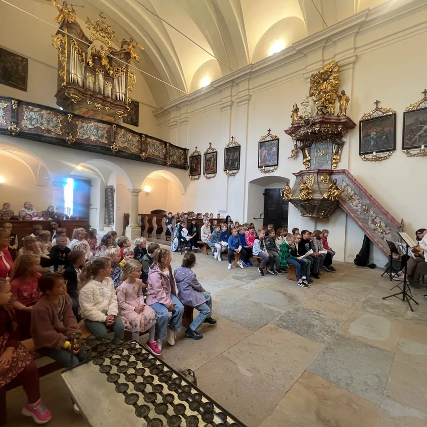 Eine Gruppe von Kindern sitzt aufmerksam in einer Kirche und hört einem Musiker zu, der auf einer großen Orgel spielt. Der Raum ist mit kunstvollen Gemälden und Holzbänken geschmückt.