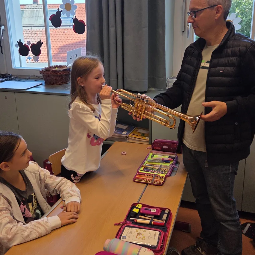 Ein junges Mädchen spielt eine Trompete, während ein erwachsener Mann sie hält, und ein anderes Mädchen beobachtet in einem Klassenzimmer.