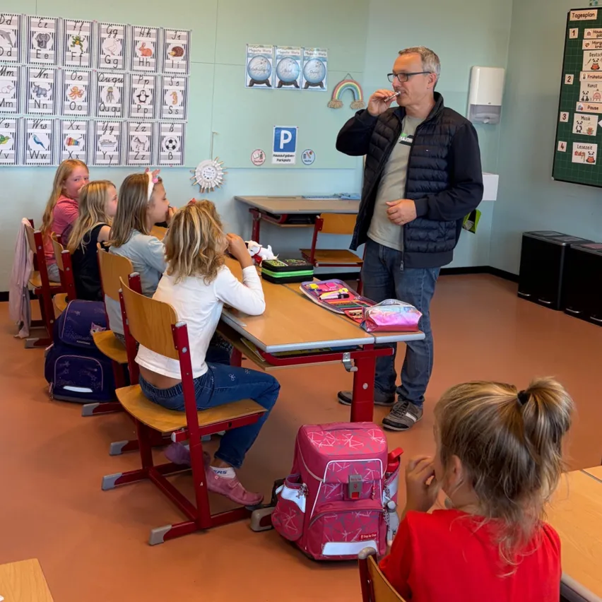 Ein Mann steht in einem Klassenzimmer und putzt seine Zähne, während junge Mädchen an ihren Tischen sitzen. Das Klassenzimmer ist mit pädagogischen Postern dekoriert.