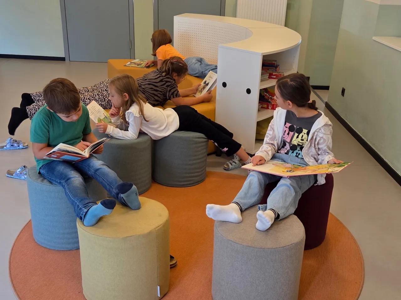 Fünf Kinder sitzen auf gepolsterten Hockern in einer Leseecke, jedes in ein Buch vertieft. Ein Mädchen liegt auf einer gelben Bank, während ein weißer Bücherregal dahinter steht.