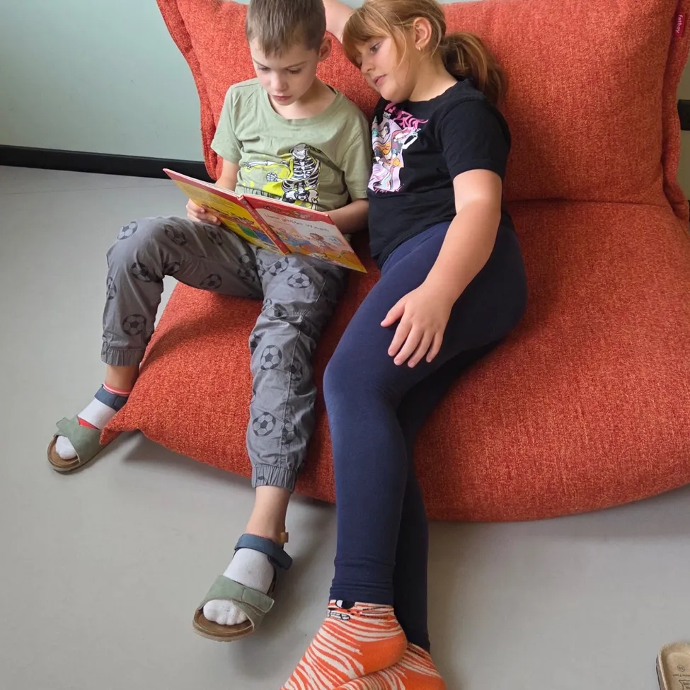 Ein Junge und ein Mädchen sitzen auf einem großen orangefarbenen Sitzsack und lesen gemeinsam ein Buch. Der Junge trägt ein grünes T-Shirt und graue Hose, während das Mädchen ein schwarzes T-Shirt und blaue Leggings trägt.