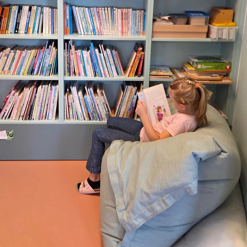 Ein kleines Mädchen sitzt in einem Sitzsack in einer Bibliothek und liest ein Buch. Die Regale hinter ihr sind mit bunten Büchern gefüllt.
