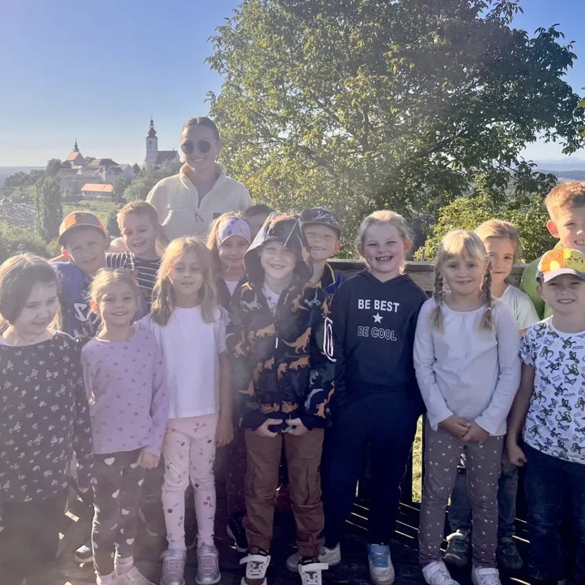 Eine Gruppe von Kindern und einem Erwachsenen stehen zusammen in einem Park, lächeln und posieren für ein Foto. Im Hintergrund sind Bäume und eine entfernte Ansicht einer Stadt zu sehen.