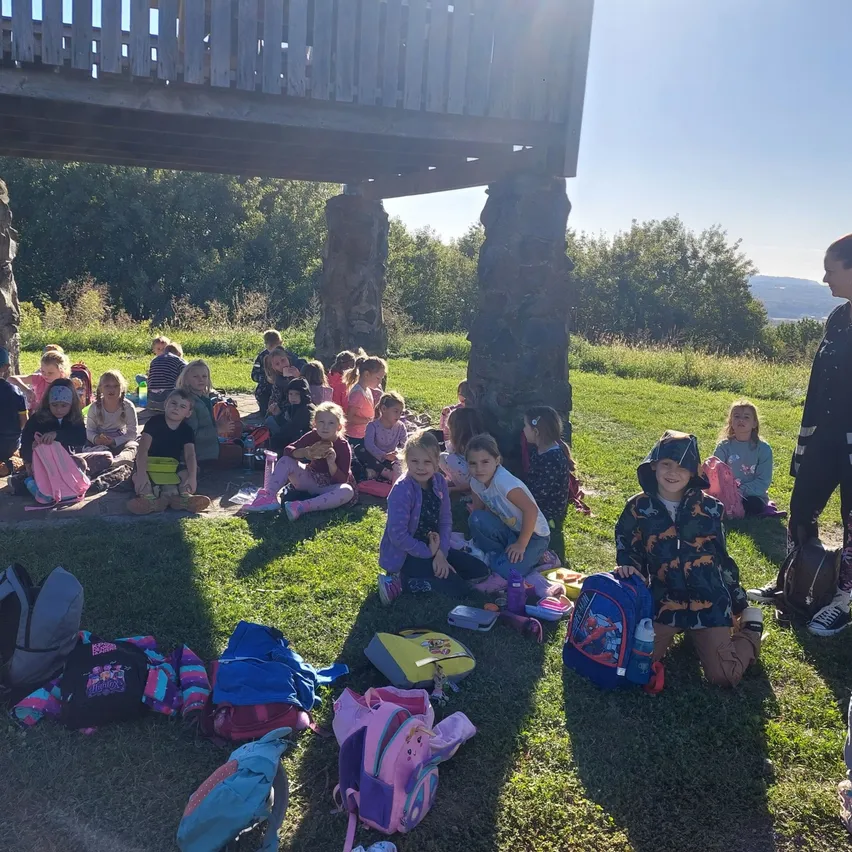 Eine Gruppe von Kindern sitzt auf dem Gras unter einer Holzstruktur, einige mit Rucksäcken und andere mit Snacks. Eine Frau steht in der Nähe.