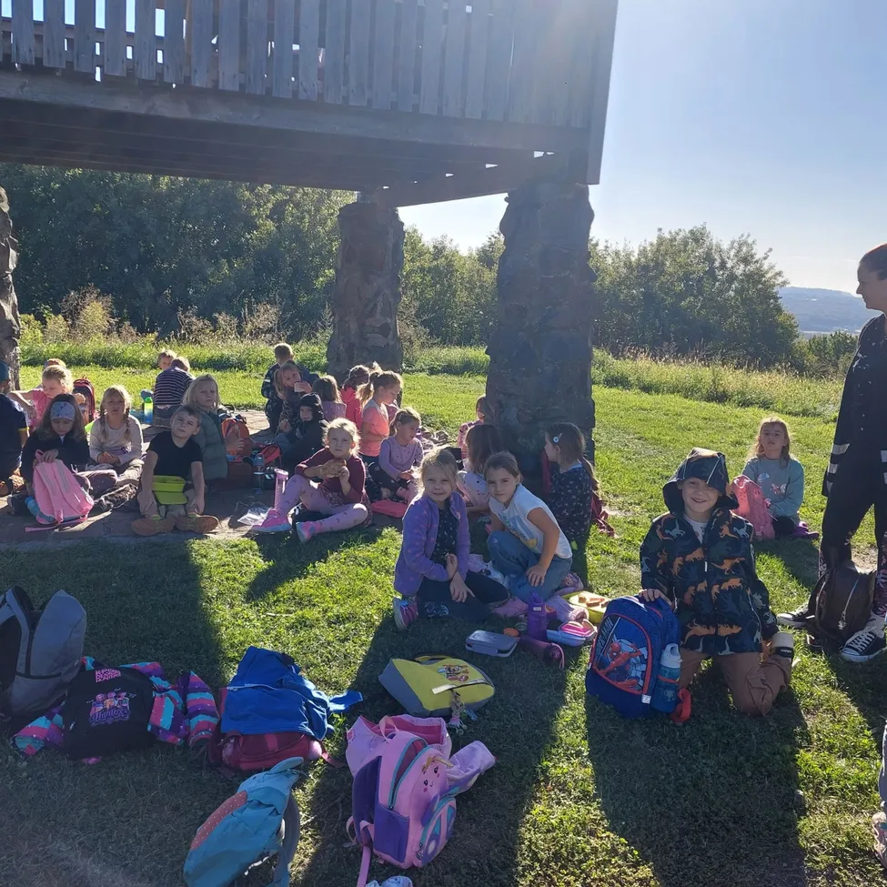 Eine Gruppe von Kindern sitzt auf dem Gras unter einer Holzstruktur, einige mit Rucksäcken und andere mit Snacks. Eine Frau steht in der Nähe.