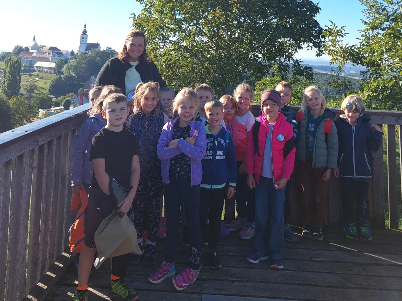Eine Gruppe von Kindern und einer Frau steht auf einer Holzplattform, lächelt und posiert für ein Foto. Sie sind von Bäumen umgeben und haben einen Blick auf eine Stadt.