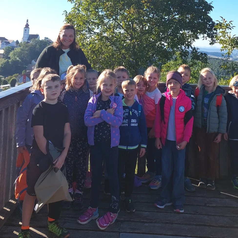 Eine Gruppe von Kindern und einer Frau steht auf einer Holzplattform, lächelt und posiert für ein Foto. Sie sind von Bäumen umgeben und haben einen Blick auf eine Stadt.