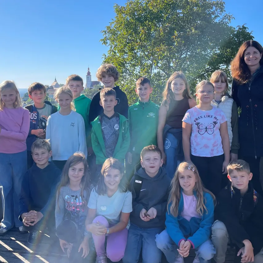 Eine Gruppe von Kindern und zwei Erwachsenen posiert für ein Foto im Freien, mit einem Baum und einem Turm im Hintergrund.
