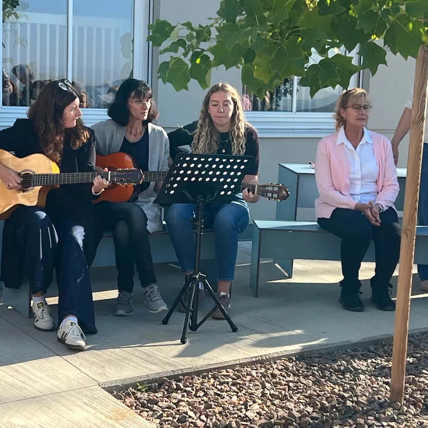 Fünf Frauen sitzen auf einer Bank vor einem Gebäude und spielen Gitarre und singen.