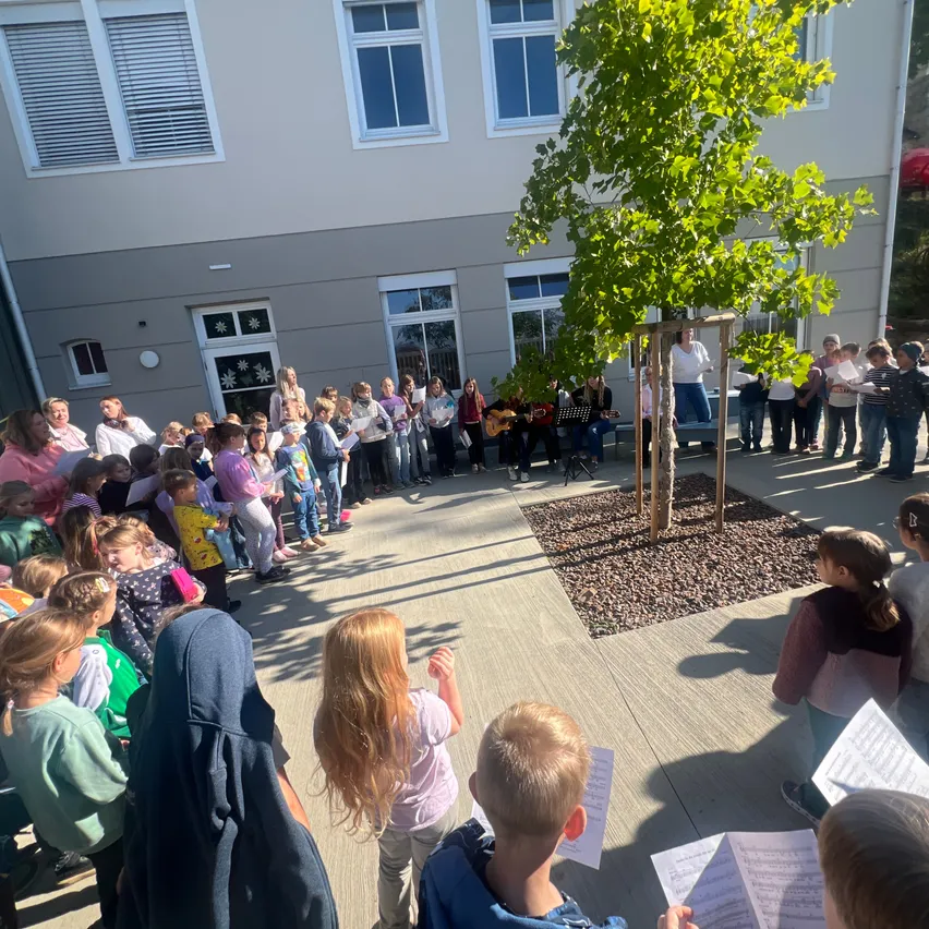 Eine Gruppe von Kindern und Erwachsenen versammelt sich auf einem Innenhof, möglicherweise für eine Schulveranstaltung. Einige halten Papiere und scheinen zu singen. Ein Baum steht in der Mitte, und ein Gebäude mit Glasfenstern ist im Hintergrund zu sehen.