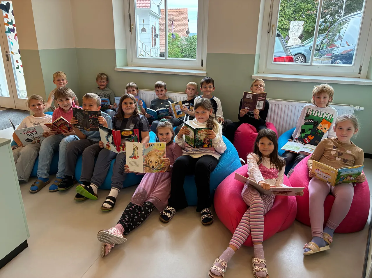 Eine Gruppe von Kindern sitzt auf Bohnensäcken und liest Bücher in einem Raum mit Fenstern und einem Heizkörper.