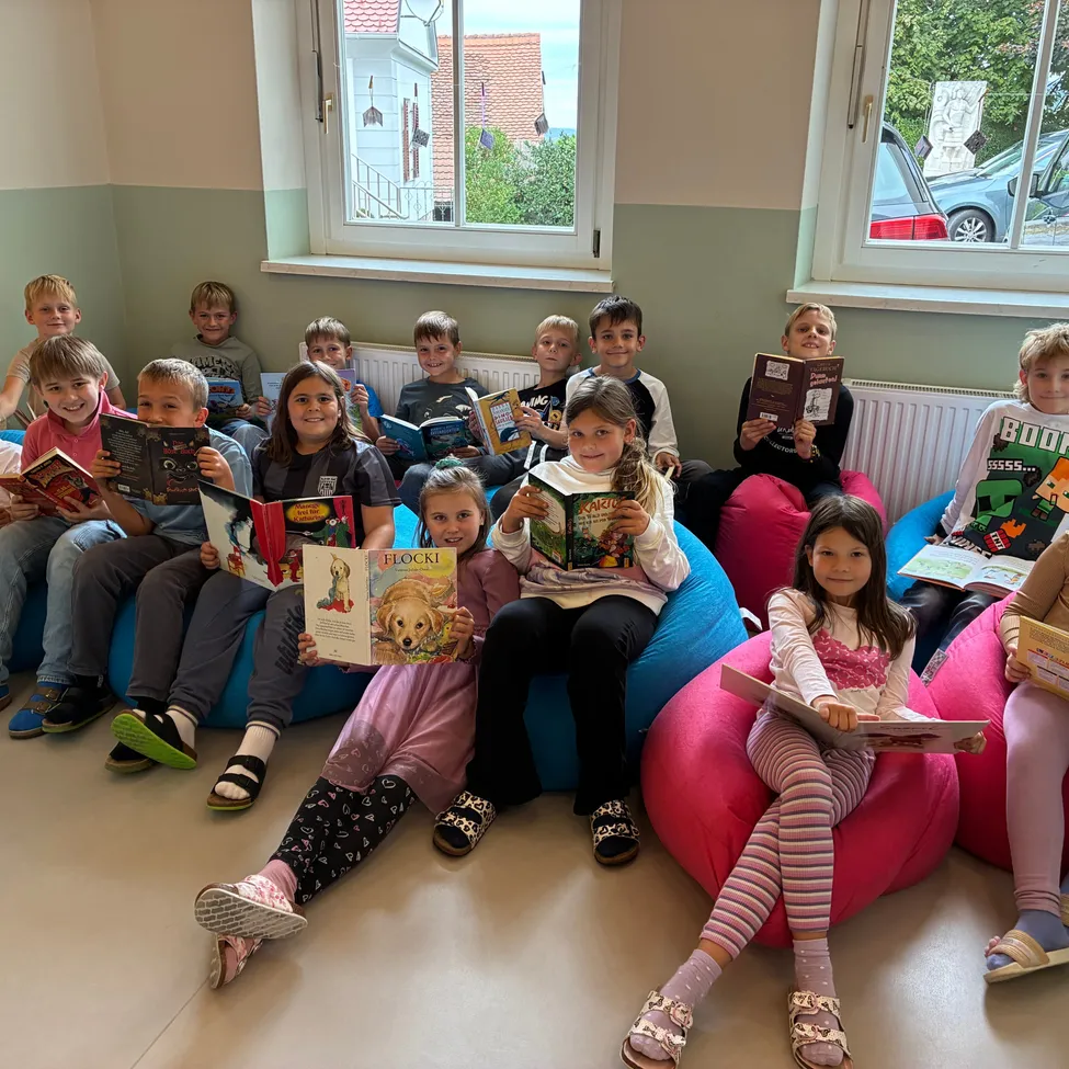Eine Gruppe von Kindern sitzt auf Bohnensäcken und liest Bücher in einem Raum mit Fenstern und einem Heizkörper.