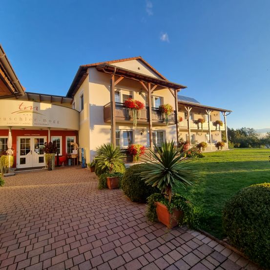 Der Eingang des Buschler-Mogge Hotels mit einem gepflasterten Weg, Topfpflanzen und einem roten Blumenbalkon an einem sonnigen Tag.