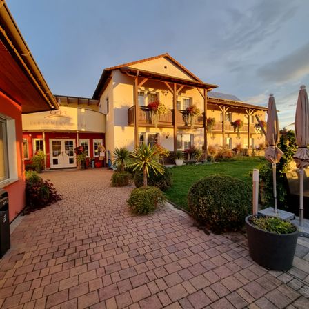 Die Vorderseite des Hotels Leuschner More in Bayern. Es hat eine rote Ziegeleinfahrt und üppige Landschaftsgestaltung.