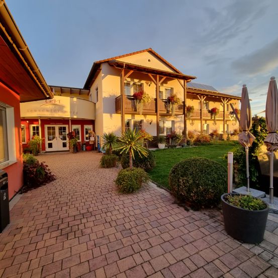 Die Vorderseite des Hotels Leuschner More in Bayern. Es hat eine rote Ziegeleinfahrt und üppige Landschaftsgestaltung.