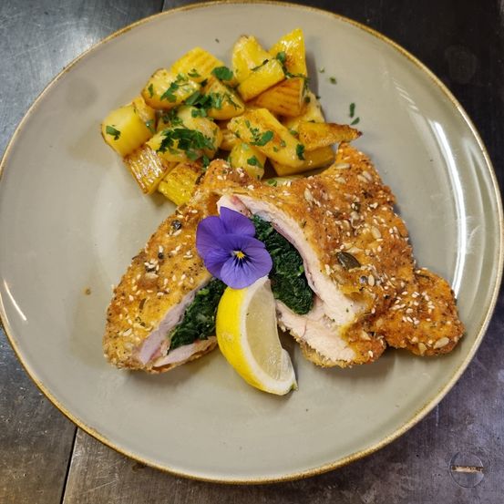 Ein Teller mit einer gebratenen Hähnchenbrust, begleitet von einer Beilage aus Kartoffelpüree und einer Zitronenscheibe, garniert mit einer lila Blume.
