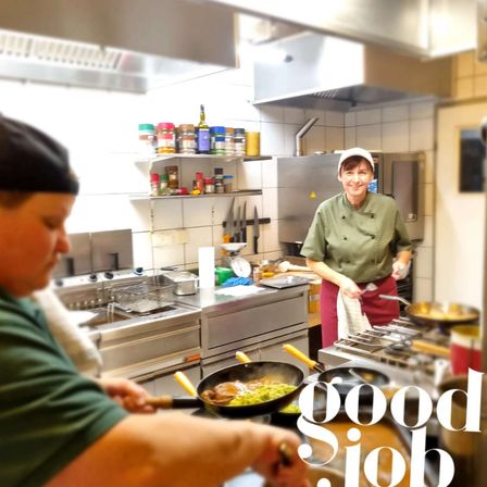 Zwei Köche in einer gewerblichen Küche, einer rührt in einem Wok und der andere lächelt, mit verschiedenen Küchenutensilien und Geräten im Hintergrund.