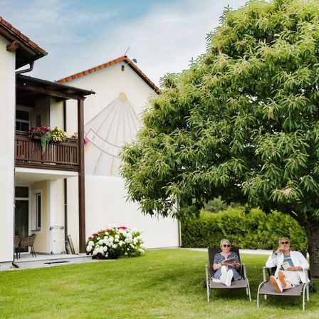Zwei Personen sitzen auf Liegestühlen unter einem Baum in einem Garten, eine liest ein Buch und die andere telefoniert. Ein Haus mit Balkon und Wandbild steht dahinter.