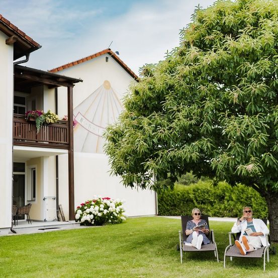 Zwei Personen sitzen auf Liegestühlen unter einem Baum in einem Garten, eine liest ein Buch und die andere telefoniert. Ein Haus mit Balkon und Wandbild steht dahinter.