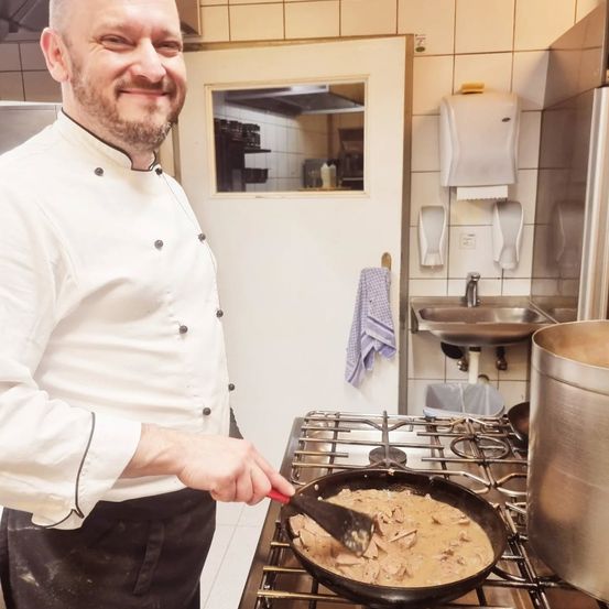 Ein Koch in weißer Uniform kocht in einer Küche mit einer Bratpfanne auf dem Herd.