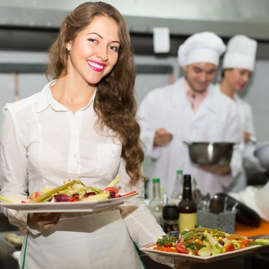 Eine Frau in einer weißen Schürze hält zwei Teller mit Salat in einer Küche, mit Köchen im Hintergrund.