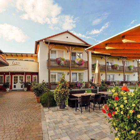 Ein Hotel mit rotem Dach und weißen Wänden, mit Balkonen mit Blumenarrangements, einer Terrasse mit Tischen und Stühlen und einer Person, die unter einem Schirm sitzt.