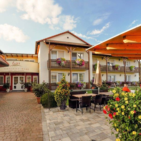 Ein Hotel mit rotem Dach und weißen Wänden, mit Balkonen mit Blumenarrangements, einer Terrasse mit Tischen und Stühlen und einer Person, die unter einem Schirm sitzt.