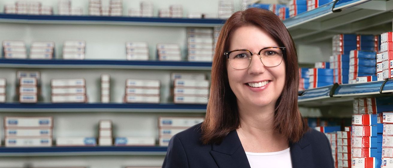 Eine lächelnde Frau in einer schwarzen Jacke und mit Brille steht vor einem Regal, das mit medizinischen Schachteln gefüllt ist.