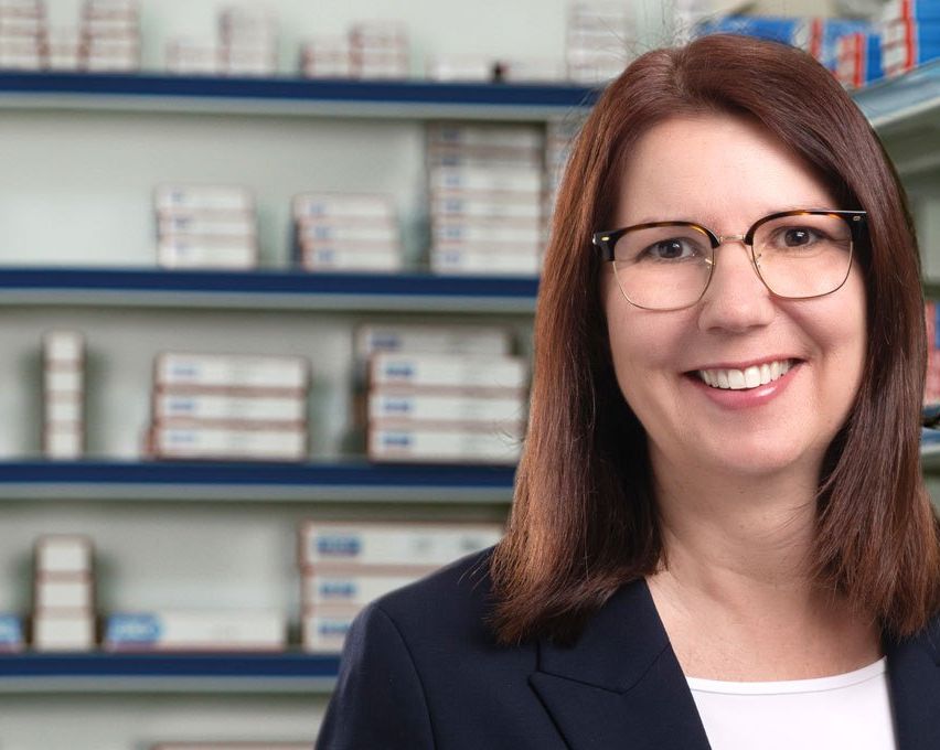 Eine lächelnde Frau in einer schwarzen Jacke und mit Brille steht vor einem Regal, das mit medizinischen Schachteln gefüllt ist.