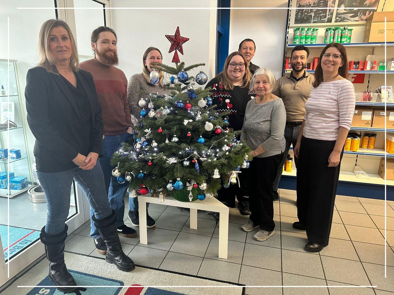 Eine Gruppe von Menschen steht um einen geschmückten Weihnachtsbaum in einem Raum. Sie alle lächeln. Eine Frau steht auf der rechten Seite.